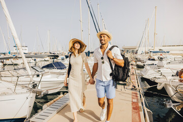 A smiling couple walks hand in hand along the marina's dock, surrounded by boats on a sunny day.
