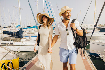 A smiling couple walks hand in hand along the marina's dock, surrounded by boats on a sunny day.
