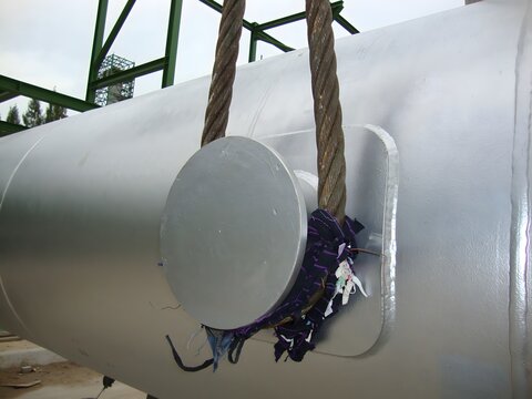 A large, cylindrical metal object, likely an industrial tank or pipe, is being lifted by thick steel cables.  A protective cloth padding is visible around the base where the cables connect.