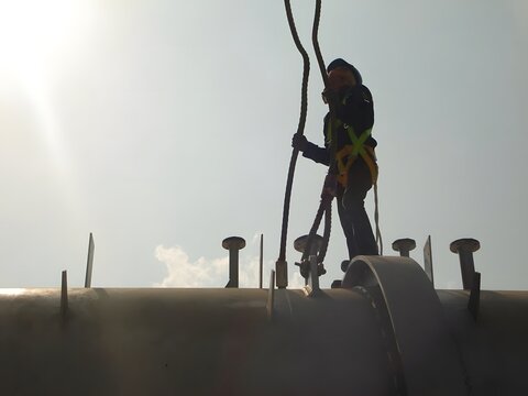 A silhouetted worker in a safety harness and helmet is perched on a large pipe, performing maintenance. The bright sky highlights the height and the critical importance of safety measures.