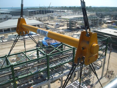 A yellow "100 TONS" spreader beam hangs from a crane over a green steel framework in an industrial construction site. Pipes, scaffolding, buildings, and the distant coastline are visible below.