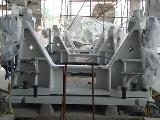 Large, white industrial molds or machinery parts are secured to a concrete base with bolts and surrounded by scaffolding and plastic sheeting, suggesting a construction or manufacturing setting.