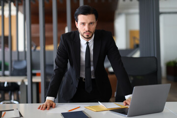 A man in a suit and tie stands in front of a desk with a laptop and a stack of papers. He is in a...