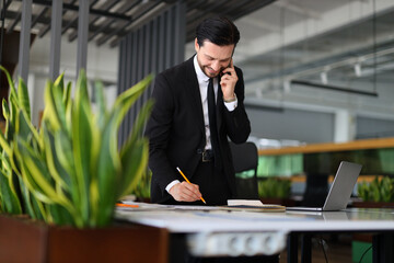 A man in a suit is talking on his cell phone while writing with a pencil. The scene suggests a busy work environment where the man is multitasking between phone calls and writing