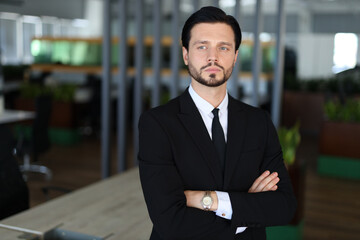 A man in a suit and tie stands in front of a desk, looking confident and professional. He is wearing a watch on his wrist, which adds to his polished appearance