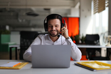 A man wearing a headset is sitting at a desk with a laptop. He is smiling and he is enjoying his...