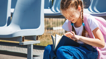 Back to school. Elementary school girl girl reads a textbook. Outdoor portrait girl with backpack on school campus