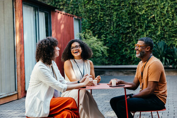 Group of coworkers enjoying coffee and laughter at an outdoor cafe setting on a sunny day,...