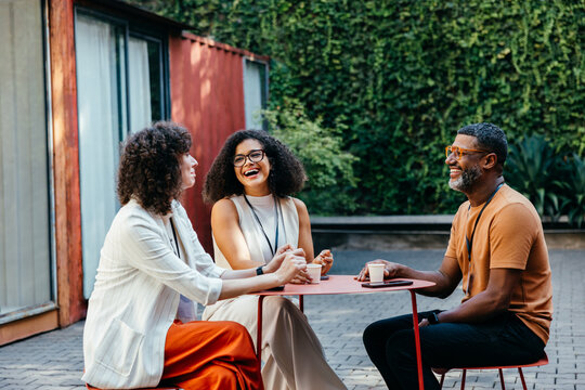 Group of coworkers enjoying coffee and laughter at an outdoor cafe setting on a sunny day, showcasing friendship and joy