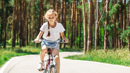 Naklejka premium Child riding bicycle. Little kid girl on bicycle along bikeway. Happy cute little girl riding bicycle. Child cycling on bicycle.