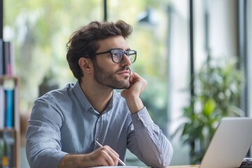 A man wearing glasses is sitting at a desk with a laptop and a pen. He is deep in thought, possibly working on a project or contemplating a decision. Concept of focus and concentration