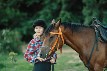 Obraz premium Happy blonde with horse in forest. Woman and a horse walking through the field during the day. Dressed in a plaid shirt and black leggings.