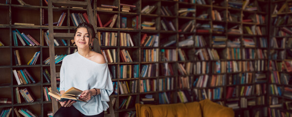 Beautiful young smiling woman holding book and looking at the camera in a home library with many...