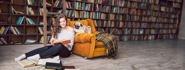 Smiling beautiful woman sitting on the floor in the library holding a book. Dog pun on orange chair reading a novel with her. Long horizontal banner. Happy relaxed weekend at home with tails