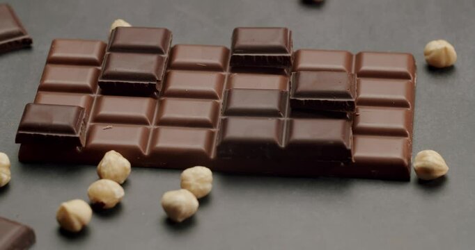 A close-up shot of a chocolate bar featuring both milk and dark chocolate pieces, surrounded by hazelnuts on a dark surface. Perfect for themes like indulgence, dessert, and confectionery.