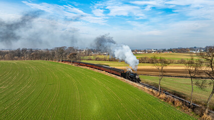 Naklejka premium A charming steam train billows white smoke as it chugs along a picturesque landscape with vibrant green fields and a clear blue sky. . High quality photo