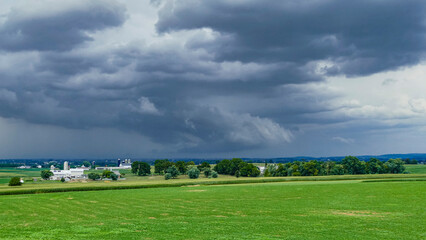 Dark clouds loom ominously over a vast green field, hinting at an approaching storm. A farm can be seen in the distance, framed by the lush countryside as day turns to dusk.