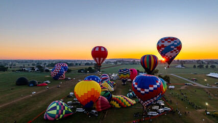 Fototapeta premium A dozen colorful hot air balloons are preparing for launch in a festival field during sunset. Spectators gather to witness the spectacle as the evening sky radiates warm colors.