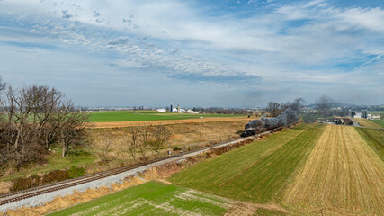 A classic steam locomotive puffs blue-gray smoke as it moves along train tracks, surrounded by lush green fields and farmland. The clear sky enhances the tranquil rural scenery.
