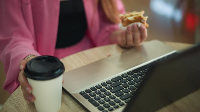 Hand view of a woman working on her laptop with a partially eaten burger in her left hand, pausing to take her drink from the table, placing it back, and continuing her work, background slightly blur