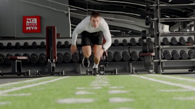A motivated male athlete bursts into a sprint on green turf within a spacious, well-equipped gym. Intensity and focus in fitness training. Focused Athlete Sprinting on Indoor Turf at a Modern Gym
