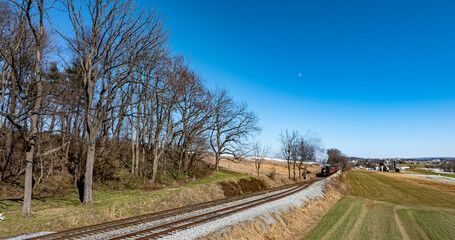 A vintage steam locomotive is making its way along the railway, surrounded by trees and rolling hills. The sky is clear and offers a stunning view of the scenery.