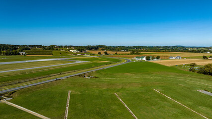 A sweeping view of lush green fields stretching towards the horizon, interspersed with winding roads and small white houses. The landscape reflects a sunny, cloudless day in the countryside.