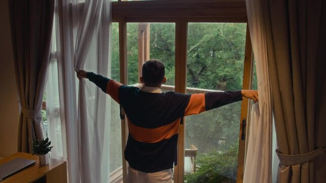 High angle view of young Caucasian man getting out of bed, opening curtains on window and stretching during morning weekend at home