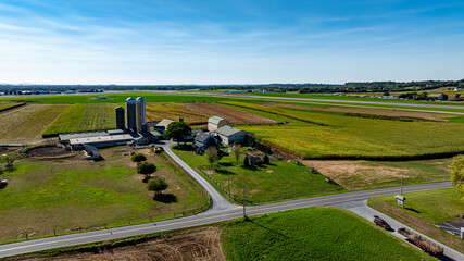 Sunny farmland stretches across the landscape, featuring green and golden fields. A farmhouse and silos stand prominently near the road, showcasing the beauty of rural agriculture.