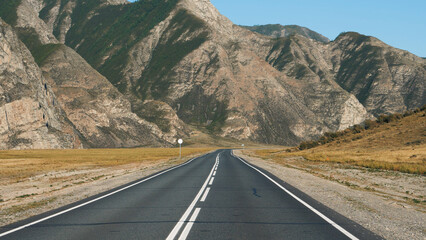 Driving on empty highway of black asphalt and white road markings at sunny day, road going into distance through desert prairie at fall to rocky mountain massive on horizon in wild. Pov moving car