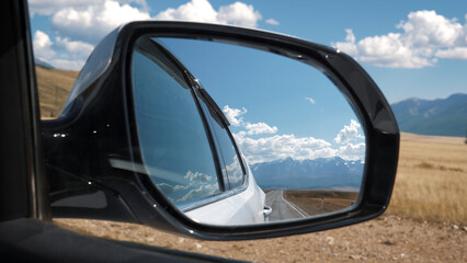 Fototapeta premium Car side rearview mirror closeup. View mountain range with snow capped summits from driving car vehicle on highway road passing through valley of foothills on sunny day. Road trip in summer holidays