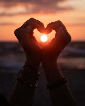 person on the beach at sunset holding and forming a heart with two hands and the sun caught inside