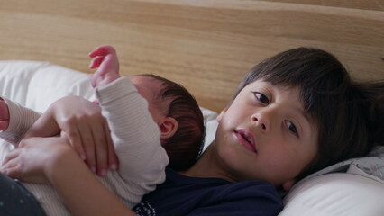 Young boy gently holding his newborn sibling on a bed, looking up at the camera with a calm expression, capturing a sweet and protective sibling moment © Marco