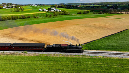 A vintage steam locomotive pulls a passenger train along a scenic route with vast green fields and patches of golden crops under a bright blue sky, creating a picturesque rural view.