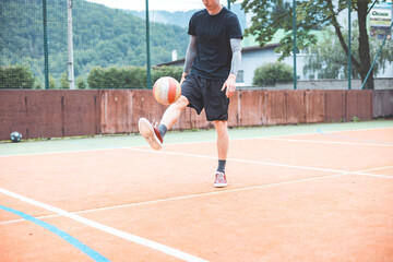 Young man casually juggles a football on an outdoor sports court, focusing on his footwork. Dressed in casual sportswear, he practices ball control in a vibrant natural setting