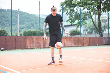 Young man casually juggles a football on an outdoor sports court, focusing on his footwork. Dressed in casual sportswear, he practices ball control in a vibrant natural setting