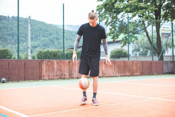 Fototapeta premium Young man casually juggles a football on an outdoor sports court, focusing on his footwork. Dressed in casual sportswear, he practices ball control in a vibrant natural setting