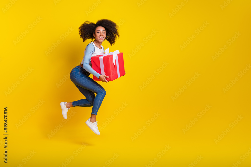 Wall mural Full body photo of attractive young woman running jump hold giftbox dressed stylish blue clothes isolated on yellow color background