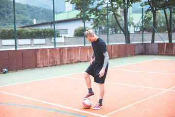 Young man with a man bun juggles a football on a vibrant orange court during a sunny day. Wearing casual sportswear, he focuses on controlling the ball with precision in a natural setting