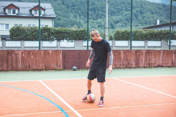 Young man with a man bun juggles a football on a vibrant orange court during a sunny day. Wearing casual sportswear, he focuses on controlling the ball with precision in a natural setting