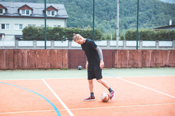 Obraz premium Young man with a man bun juggles a football on a vibrant orange court during a sunny day. Wearing casual sportswear, he focuses on controlling the ball with precision in a natural setting