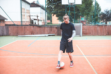 Obraz premium Young man with a man bun juggles a football on a vibrant orange court during a sunny day. Wearing casual sportswear, he focuses on controlling the ball with precision in a natural setting