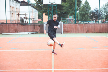 Obraz premium Young man with a man bun juggles a football on a vibrant orange court during a sunny day. Wearing casual sportswear, he focuses on controlling the ball with precision in a natural setting