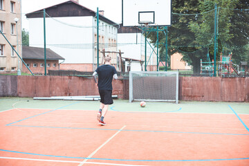 Fototapeta premium Young man runs towards the goal, preparing to shoot a football. The outdoor court features an urban backdrop with buildings and a basketball hoop, combining casual sports with focus and energy