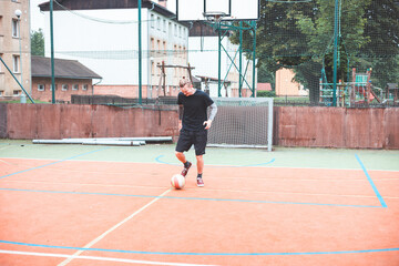 Fototapeta premium Young man focuses on controlling a football on an outdoor court. He carefully juggles the ball with precision, surrounded by an urban backdrop of buildings and a goal, combining sports and skill