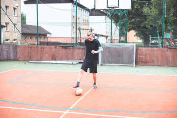 Young man focuses on controlling a football on an outdoor court. He carefully juggles the ball with precision, surrounded by an urban backdrop of buildings and a goal, combining sports and skill