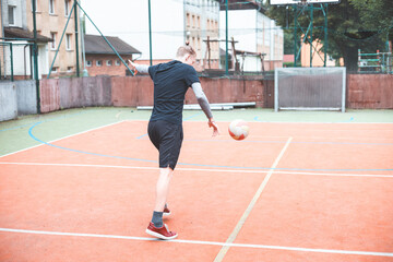 Fototapeta premium Young man kicks a football on an outdoor court, balancing his posture for a powerful shot. Wearing sporty attire, he plays in a vibrant setting with a goal and basketball hoop behind him