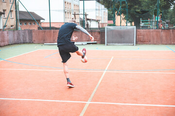 Obraz premium Young man kicks a football on an outdoor court, balancing his posture for a powerful shot. Wearing sporty attire, he plays in a vibrant setting with a goal and basketball hoop behind him