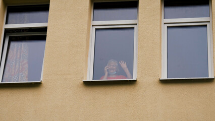 Elderly old woman sick coronavirus infection, stands at window in hospital and communicates through glass on phone with relatives and family, waving hand greeting. Isolation during covid-19 pandemic