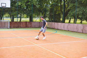 Obraz premium Young man dribbles a football with precision on an outdoor court, practicing his footwork in a casual environment. The court is surrounded by nature, providing a peaceful backdrop